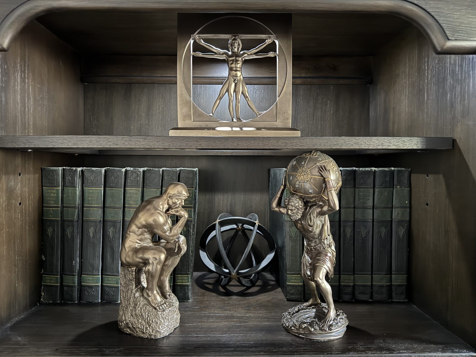 A bookcase: a bronze Vitruvian Man above; below, Rodin's Thinker, an armillary sphere and Atlas carrying the celestial globe, between volumes of Compton's Pictured Encyclopedia